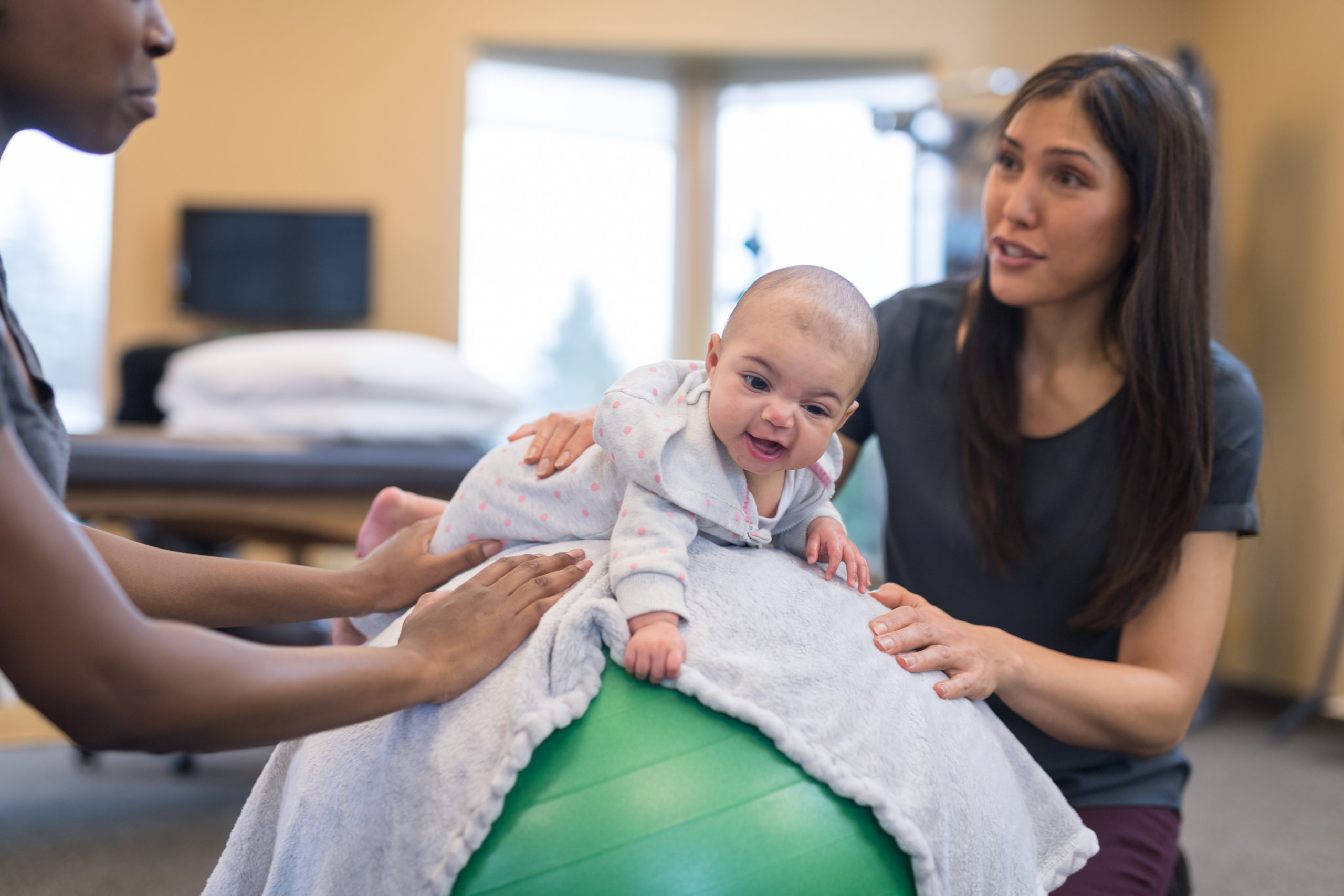 A physical therapist of Asian descent works with an infant. She and baby's mom are helping her lie face down on an exercise ball while she practices balancing.